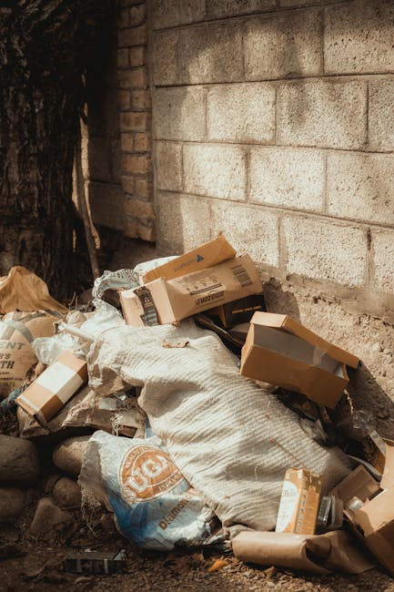 A pile of mixed waste materials located against an exterior brick and concrete wall, including flattened cardboard boxes, partially filled paper bags, and crumpled packaging. The cardboard boxes vary in size, with some open and others still folded, displaying plain brown surfaces with printed labels. The paper bags are beige and appear to be made of plain paper, with some torn or crumpled. Large woven white sacks contain additional waste, with some parts loosely spilling onto the ground, which is uneven and composed of dark soil and small rocks. In the background, a concrete wall with visible mortar joints and a brick section are partly obscured by a large tree trunk on the left side of the image. The scene is outdoors, illuminated by natural daylight, with the waste positioned directly adjacent to the wall, suggesting possible on-site clearance or private disposal handled by companies like Rubbish Removal Hampstead, providing an example of non-collective waste management or alternative rubbish removal services.