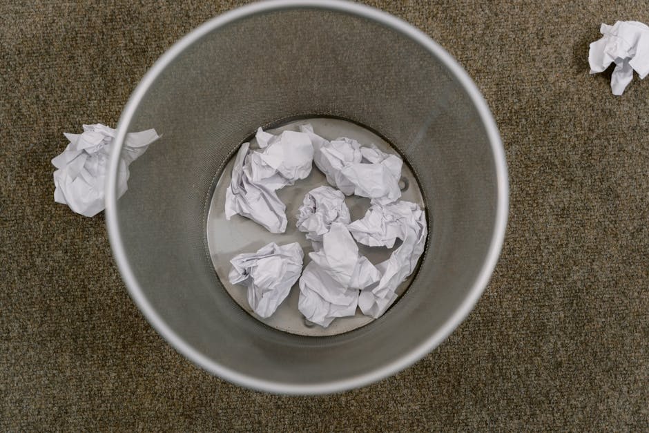A round, metallic rubbish bin with a smooth, matte finish, placed on a textured, brown carpeted floor. Inside the bin, several crumpled sheets of white paper are visible, some partially submerged in water, indicating dampness or recent disposal. Around the bin, additional crumpled paper scraps are scattered on the floor, suggesting a messy disposal area. The lighting is neutral and evenly distributed, with no shadows or reflections. The scene appears to be part of an indoor office or home environment, emphasizing the importance of proper waste management and alternative rubbish disposal methods, such as on-site clearance or private collection services provided by Rubbish Removal Hampstead. The overall composition highlights the practical aspects of rubbish removal, focusing on the collection and handling of crumpled paper waste in a straightforward, factual manner.