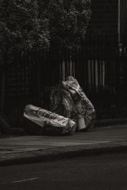 A black and white image showing two large, crumpled black plastic bags filled with waste resting on the edge of a paved sidewalk next to a dark wooden fence with vertical slats. The bags appear to contain bulky rubbish and are positioned side by side, partially leaning against the fence. In the background, dense foliage from a tree is visible, providing some natural contrast behind the fencing. The scene is illuminated by natural light, emphasizing the textures of the plastic bags and the surface of the pavement. The setting suggests an outdoor area requiring further clearance or collection, consistent with private waste removal services such as those provided by Rubbish Removal Hampstead, offering an alternative to municipal rubbish collection for difficult access locations or larger waste items.