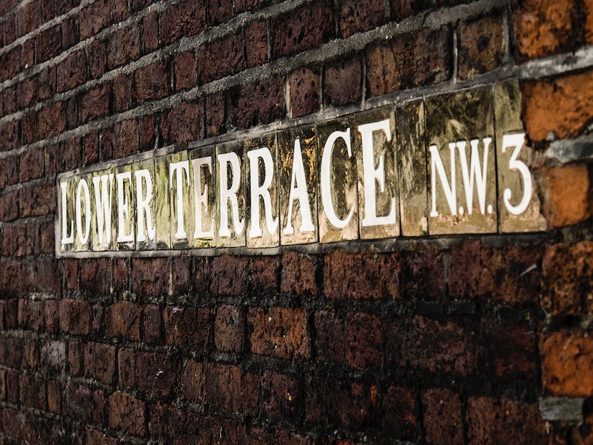 A close-up image of a horizontal street sign mounted on a weathered brick wall. The sign reads 'LOWER TERRACE N.W.3' in bold, white uppercase letters on a reflective metallic background with some visible weathering and moss growth, indicating age and exposure to the elements. The brick wall behind the sign consists of reddish-brown bricks with dark mortar, featuring a rough texture and slightly uneven surface. The overall scene suggests an urban setting, possibly in a residential or service area, where the sign provides directional or location information. The lighting is natural, highlighting the textures and contrasts between the shiny sign and the matte brick surroundings, subtly reflecting the environment connected to private waste handling or alternative rubbish collection methods often associated with on-site clearance services by companies like Rubbish Removal Hampstead.