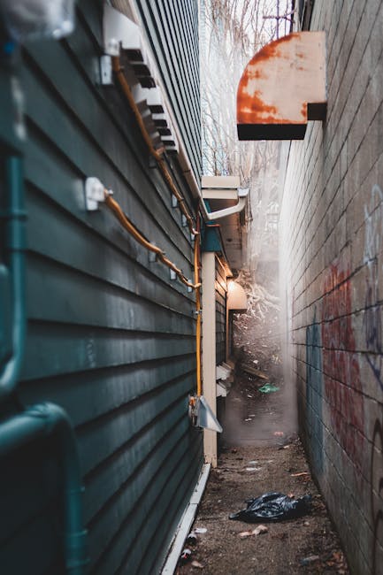 A narrow outdoor alleyway between two buildings, with the left side featuring dark green horizontal corrugated metal cladding and visible orange gas pipes attached to the wall. The right side consists of a tall, weathered concrete or brick wall with some graffiti. The ground is uneven, muddy, and littered with scattered debris, including a black plastic bag near the foreground. In the background, there are faint trees and a cloudy sky, with diffused natural light illuminating the scene. A small, mounted outdoor light fixture is attached to the wall on the right, casting a soft glow. The alley appears to be an area accessible for waste removal or private clearance, with signs of neglect and accumulated refuse, consistent with a setting where independent rubbish collection services, such as those offered by Rubbish Removal Hampstead, might operate to clear clutter in tight access locations.