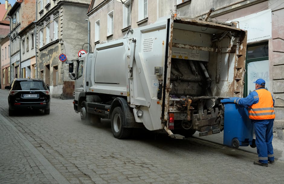 A narrow outdoor alleyway between two buildings, with the left side featuring dark green horizontal corrugated metal cladding and visible orange gas pipes attached to the wall. The right side consists of a tall, weathered concrete or brick wall with some graffiti. The ground is uneven, muddy, and littered with scattered debris, including a black plastic bag near the foreground. In the background, there are faint trees and a cloudy sky, with diffused natural light illuminating the scene. A small, mounted outdoor light fixture is attached to the wall on the right, casting a soft glow. The alley appears to be an area accessible for waste removal or private clearance, with signs of neglect and accumulated refuse, consistent with a setting where independent rubbish collection services, such as those offered by Rubbish Removal Hampstead, might operate to clear clutter in tight access locations.