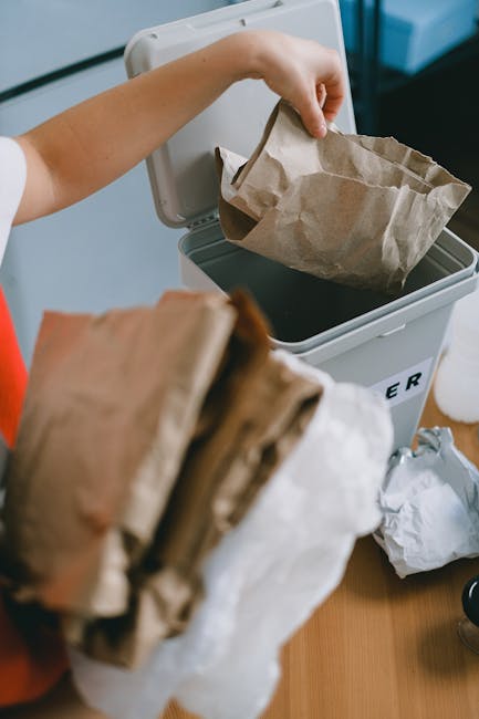 A person is disposing of a crumpled brown paper bag into a white rubbish bin with a black interior. The person's hand is visible holding the bag, and they are wearing a red sleeve. The bin is positioned on a wooden surface, likely a table or countertop. Surrounding the bin are additional paper waste items, including more crumpled paper bags and sheets, indicative of small waste quantities typically associated with small business rubbish. The environment appears to be an indoor space, away from public trash collection points, suggesting the scene relates to private or independent waste disposal methods such as on-site rubbish management by services like Rubbish Removal Hampstead. The scene emphasizes the practical aspect of rubbish handling, with standard waste materials and a focus on organized waste separation, relevant to businesses seeking alternative waste collection solutions.