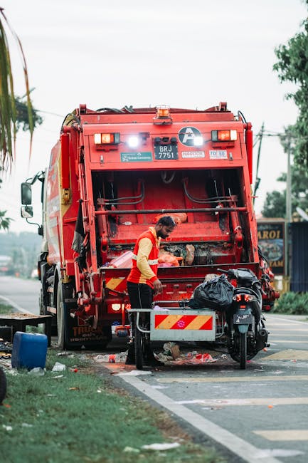 A street scene showing a waste collection worker in a red and yellow uniform operating a large red rubbish truck parked at the edge of the pavement. The rear of the truck, with its various mechanical components and safety lights, is prominently visible. The worker is standing in front of the open bin area, handling a black plastic bag filled with waste. To the side of the truck, a small motorcycle is parked partially blocking a recycling or waste collection container. The surrounding environment includes a paved road, a grassy verge with some small plants, and a few trees in the background under an overcast sky. The scene suggests an on-site waste disposal or rubbish removal process, typical of private or independent collections that avoid municipal rubbish services. Nearby, there is a glimpse of a building with signage, and the overall scene emphasizes a professional, neutral approach to waste management activities, consistent with services like those offered by Rubbish Removal Hampstead.