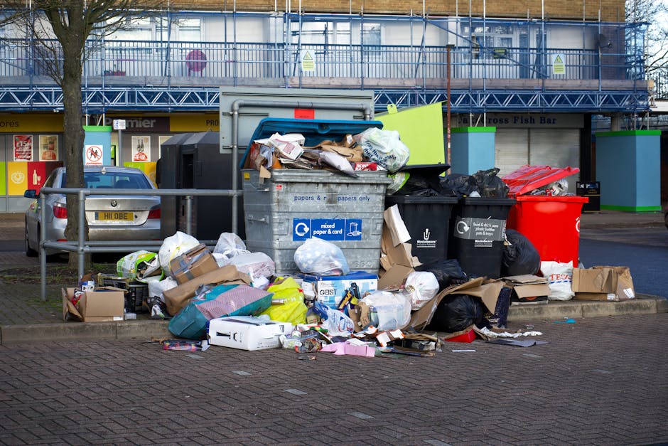 A large pile of mixed domestic waste and refuse bags is collected around several litter bins on a paved urban street near a commercial area. The waste includes crumpled cardboard boxes, plastic packaging, paper, and black and red garbage bags, some of which are torn or overflowing, spilling rubbish onto the ground. Behind the waste collection points, there is a parked silver car with a visible license plate, and a metal barrier partially surrounds the area. In the background, there is a building with blue scaffolding and a glass-fenced balcony, indicating ongoing construction or maintenance work. Adjacent to the waste area, shopfronts are visible, including a fish bar and an entrance with signage, suggesting a high foot traffic location where independent rubbish collection may be necessary for efficient waste management, aligning with services offered by Rubbish Removal Hampstead. The scene portrays a typical urban waste collection spot, emphasizing the importance of proper waste disposal and removal within the local community environment.