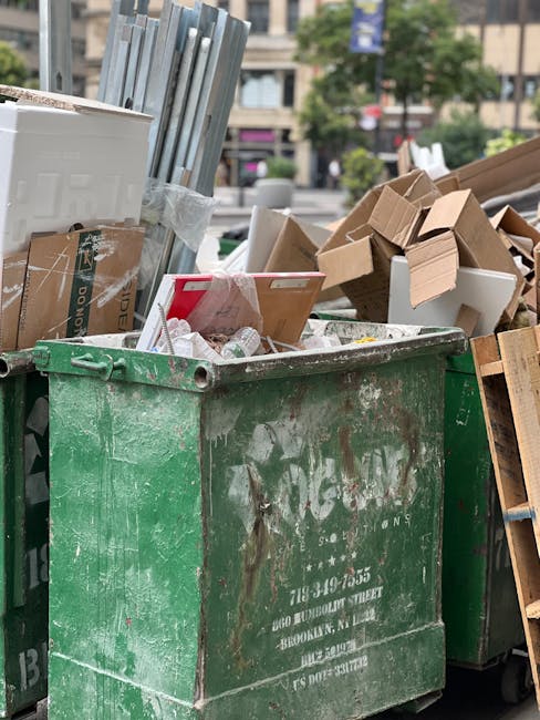 A large, open green waste bin filled with various discarded materials, including plastic bottles and paper, sits on a pavement in an urban environment. Surrounding the bin are piles of flattened cardboard boxes, some partially torn or crumpled, and a collection of wooden items such as a slatted wooden panel leaning against the bin. The background features a city street scene with blurred buildings, trees, and parked vehicles, indicating a busy area. The bin appears weathered with visible dirt and rust, characteristic of frequently used waste collection containers. Natural daylight illuminates the scene, highlighting the different textures of the materials and the slight grime on the bin's surface. This scene reflects a typical setting for independent waste removal services, such as those provided by Rubbish Removal Hampstead, handling bulky or mixed waste outside residential or commercial premises. The arrangement emphasizes the importance of proper waste management and disposal in urban environments, supporting alternative waste handling methods apart from council-operated collections.