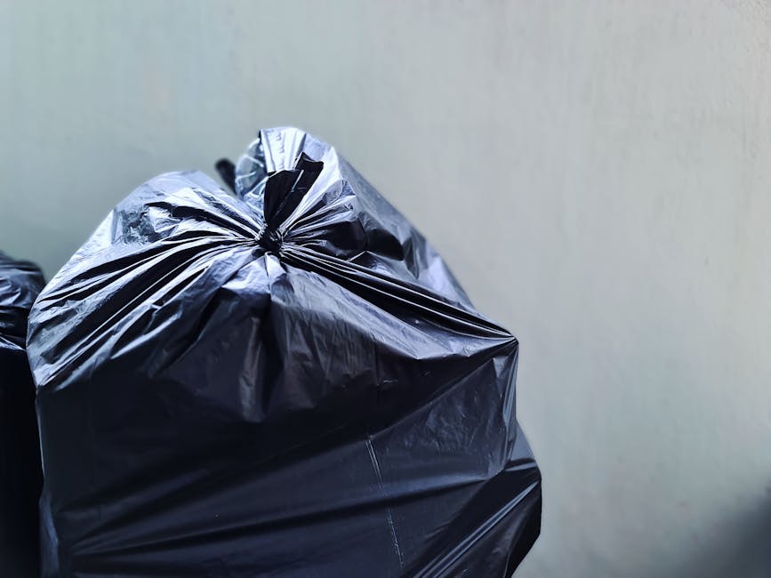 Close-up of multiple black plastic rubbish bags filled with household waste, tied at the top with knots, stacked against a plain, light-colored wall. The bags have a glossy finish and appear to be of heavy-duty material suitable for waste collection. The background shows a neutral-toned surface, possibly a wall or a corner of an indoor or outdoor storage area. The bags are positioned in the foreground, with one partially overlapping others, suggesting they are ready for collection or disposal, which aligns with private rubbish removal services provided by companies like Rubbish Removal Hampstead. The environment appears clean, without visible debris or additional objects, emphasizing the focus on waste storage or on-site clearance. The lighting is even, with no harsh shadows, highlighting the texture of the plastic and the form of the stacked bags. This scene exemplifies typical private waste management practices, supporting the concept of independent rubbish removal as an alternative to local authority collection services.