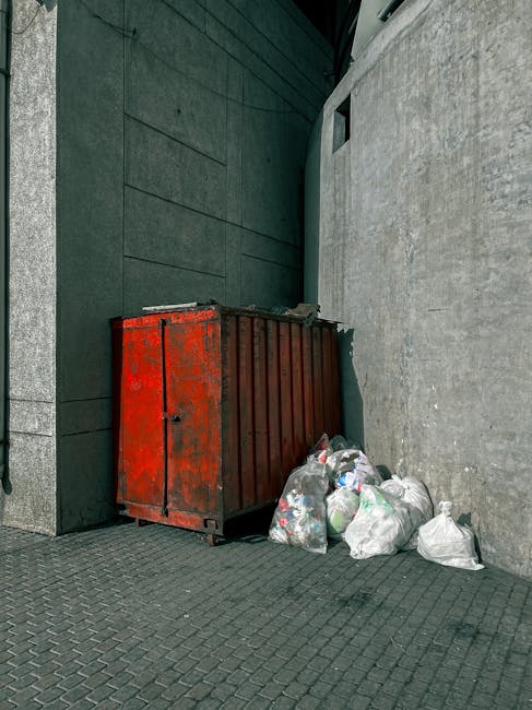 Close-up of multiple black plastic rubbish bags filled with household waste, tied at the top with knots, stacked against a plain, light-colored wall. The bags have a glossy finish and appear to be of heavy-duty material suitable for waste collection. The background shows a neutral-toned surface, possibly a wall or a corner of an indoor or outdoor storage area. The bags are positioned in the foreground, with one partially overlapping others, suggesting they are ready for collection or disposal, which aligns with private rubbish removal services provided by companies like Rubbish Removal Hampstead. The environment appears clean, without visible debris or additional objects, emphasizing the focus on waste storage or on-site clearance. The lighting is even, with no harsh shadows, highlighting the texture of the plastic and the form of the stacked bags. This scene exemplifies typical private waste management practices, supporting the concept of independent rubbish removal as an alternative to local authority collection services.